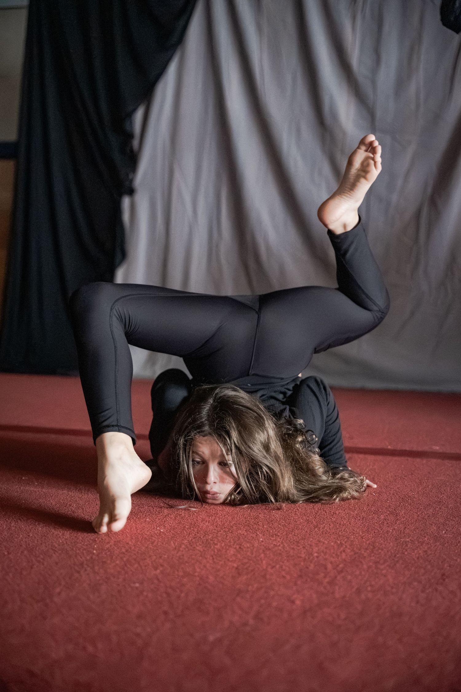 Flexible contortionist in black outfit performing advanced floor pose with legs overhead on red carpet.