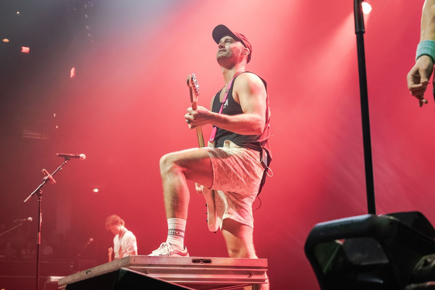 Male performer in red shorts and cap stands on stage platform holding microphone under dramatic red stage lighting.