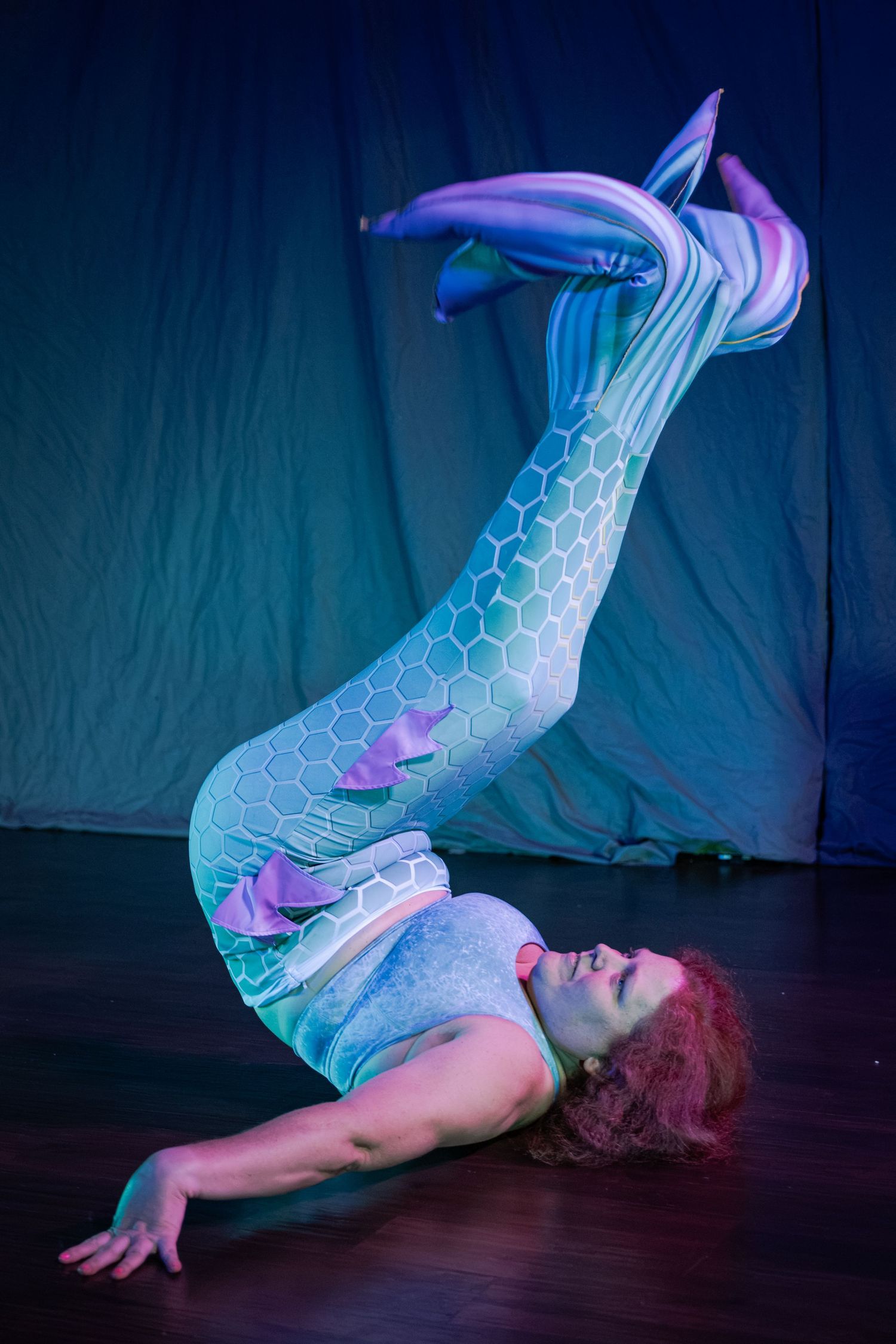 Performer in mermaid costume doing acrobatic floor pose under blue and purple stage lighting.