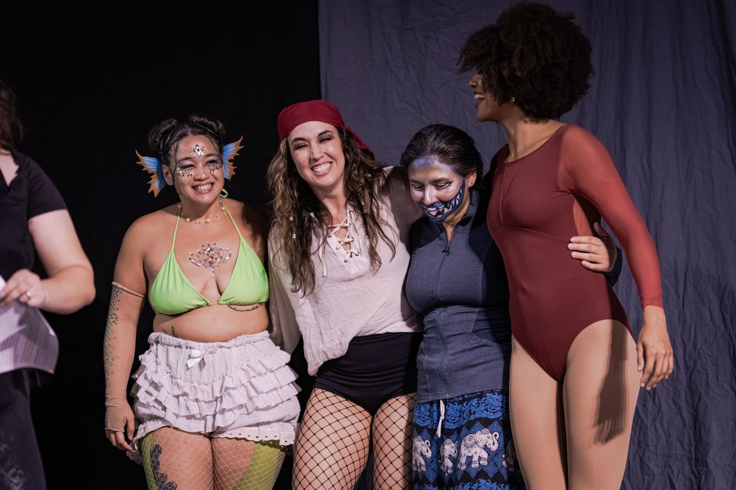 Four women posing together smiling at an indoor event, wearing colorful costumes including a green bikini top and red beret.