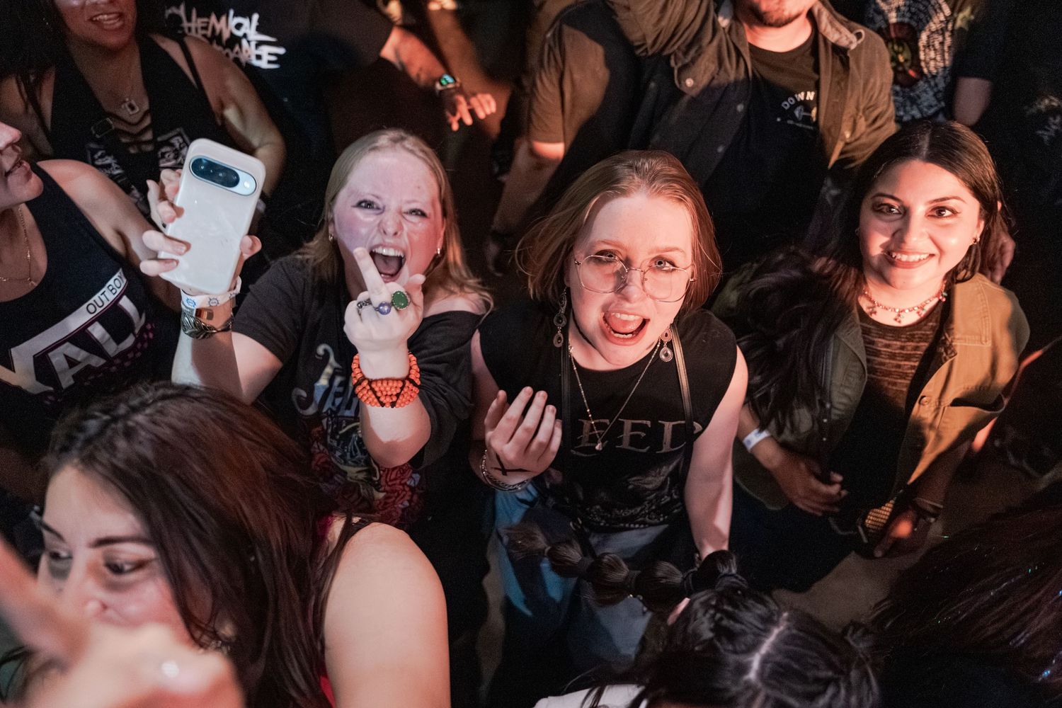 Excited fans at a rock concert crowd, women smiling and making faces, one holding a phone for a selfie.