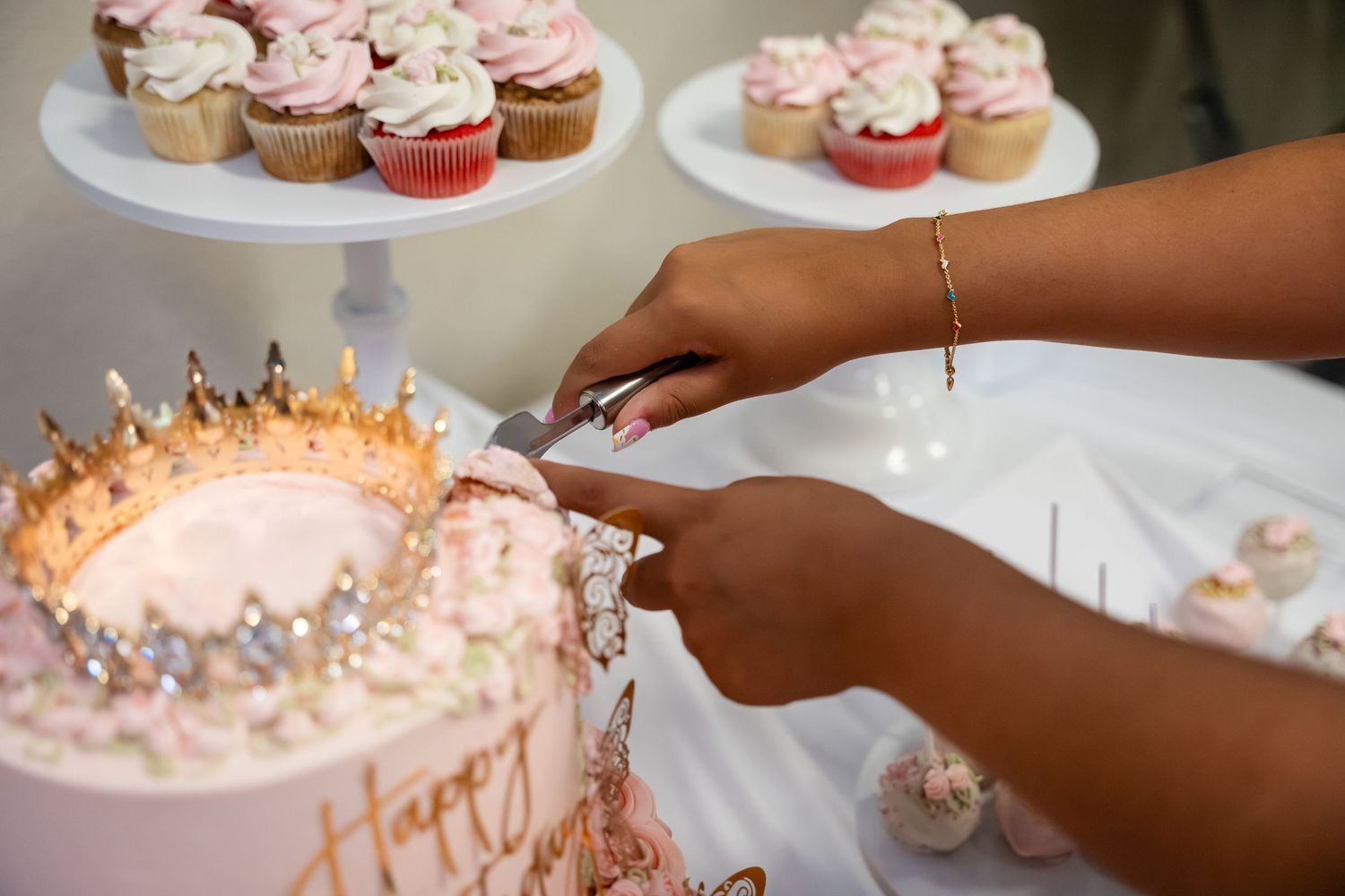 Person cutting a decorated pink birthday cake with gold crown, cupcakes on stand in background.