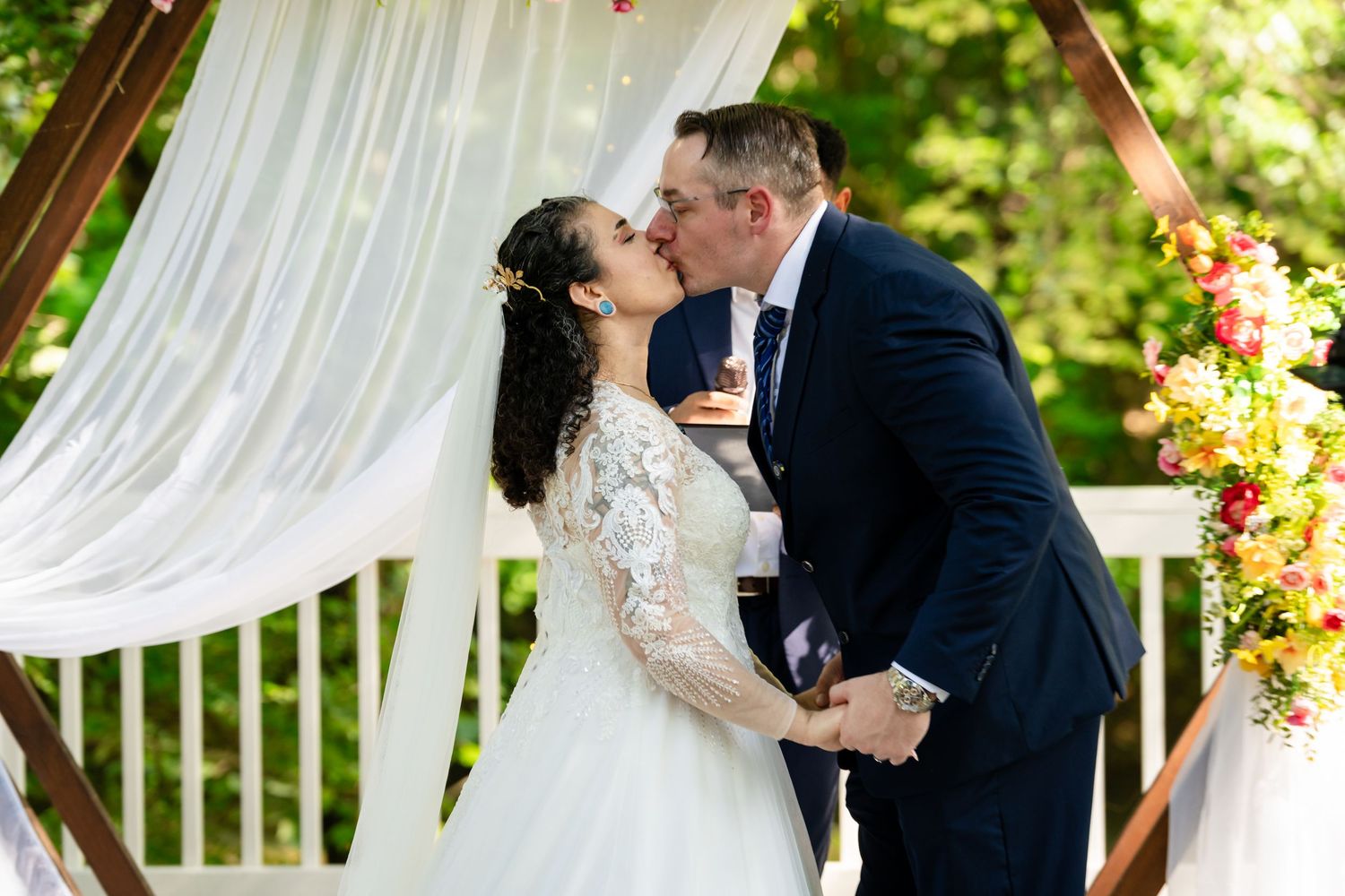 Bride and groom share first kiss at outdoor wedding ceremony under white draped arch with colorful floral arrangements.