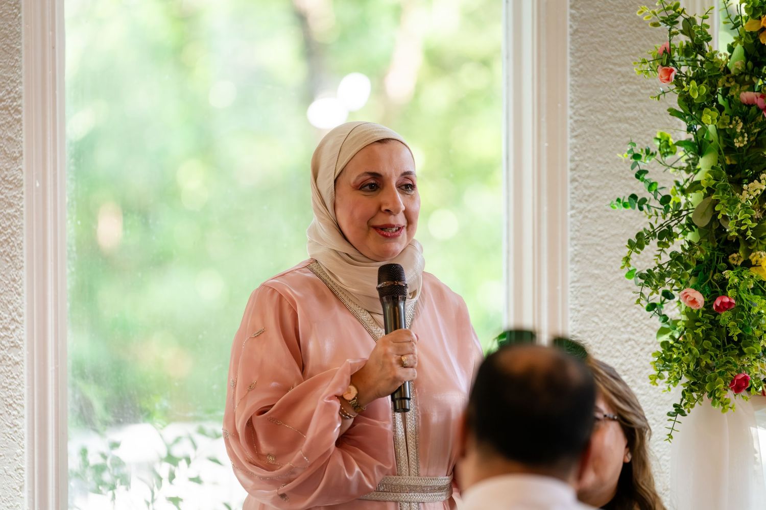 Woman in pink hijab speaking into microphone at elegant indoor event with floral decorations.