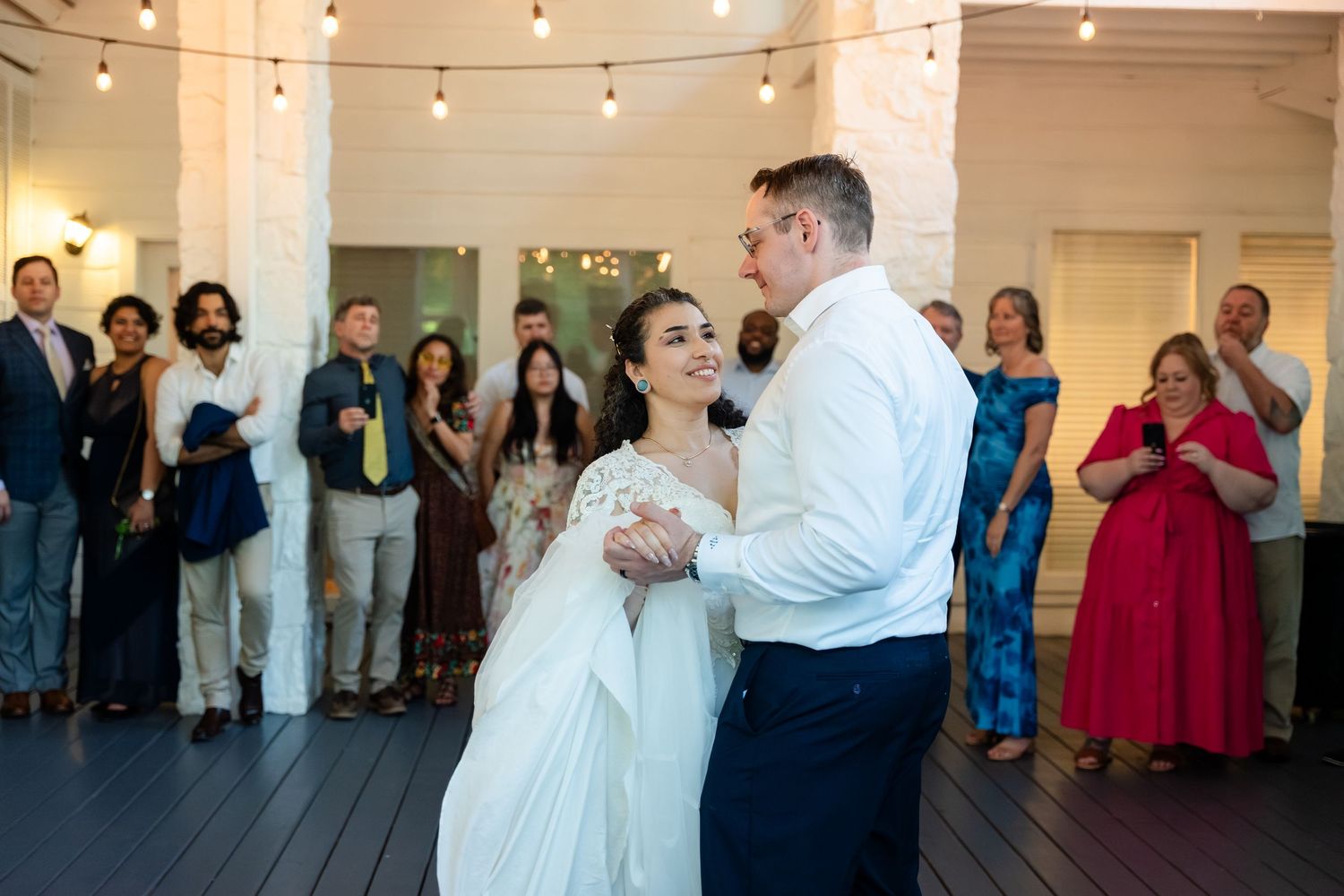Bride and groom share their first dance at an outdoor wedding reception with guests watching under string lights.