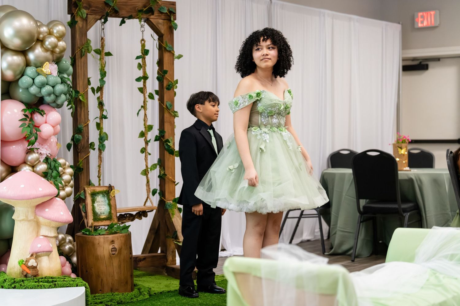 Girl in green dress and boy in tuxedo pose at elegant fairy tale themed party with mushroom decor and balloon arch.