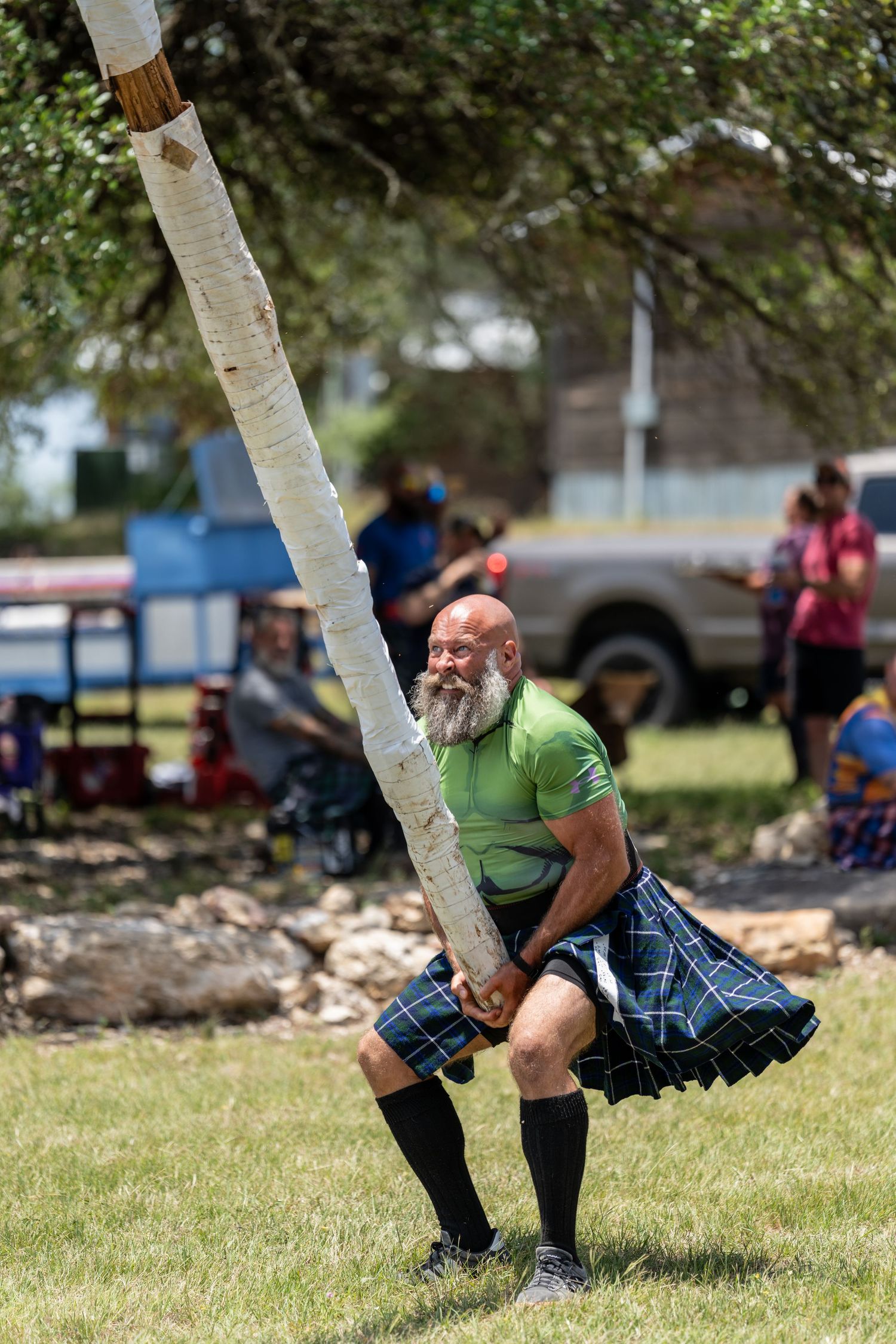 Muscular bald man with beard wearing kilt tossing caber at Scottish Highland Games competition outdoors.