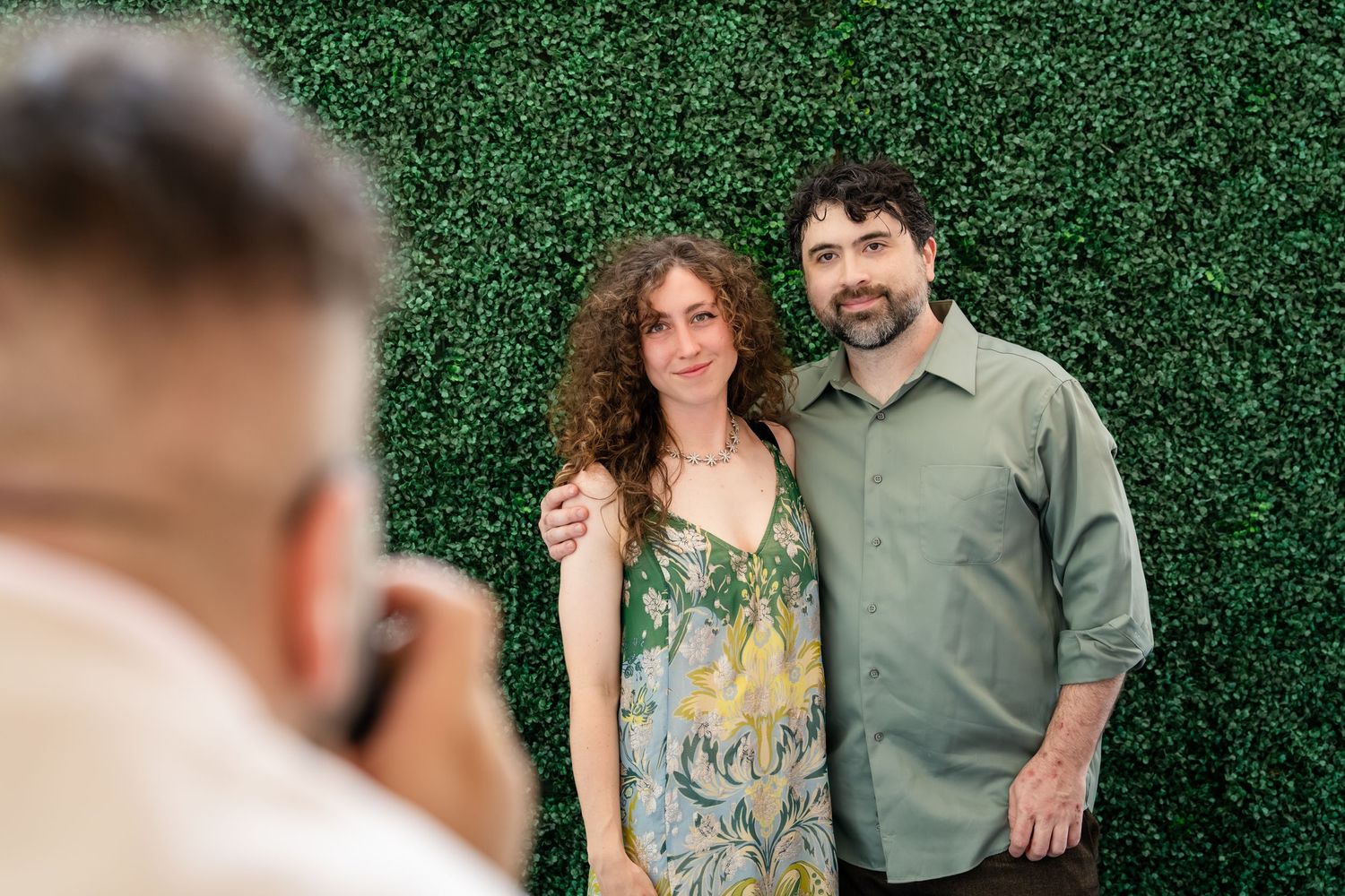 A couple poses smiling in front of a green hedge wall while a photographer captures their portrait outdoors.
