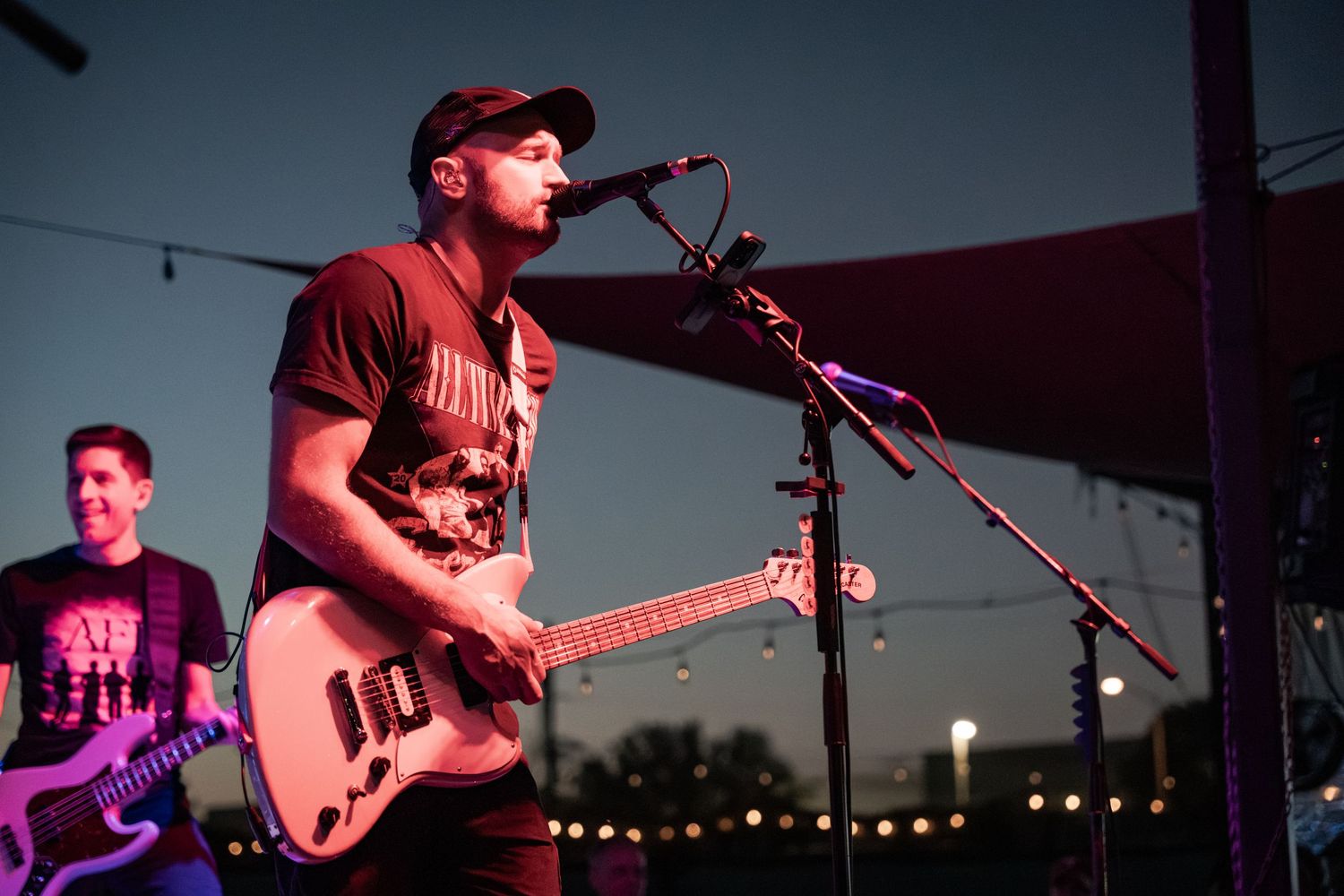 Male guitarist singing into microphone on outdoor stage at dusk during live music performance.