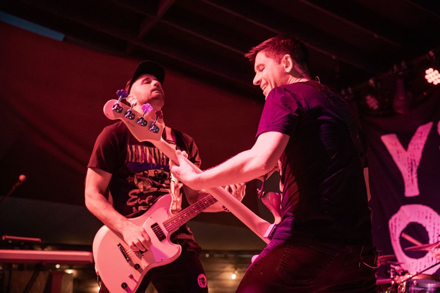 Two guitarists performing energetically on stage under pink lights at a live rock concert.