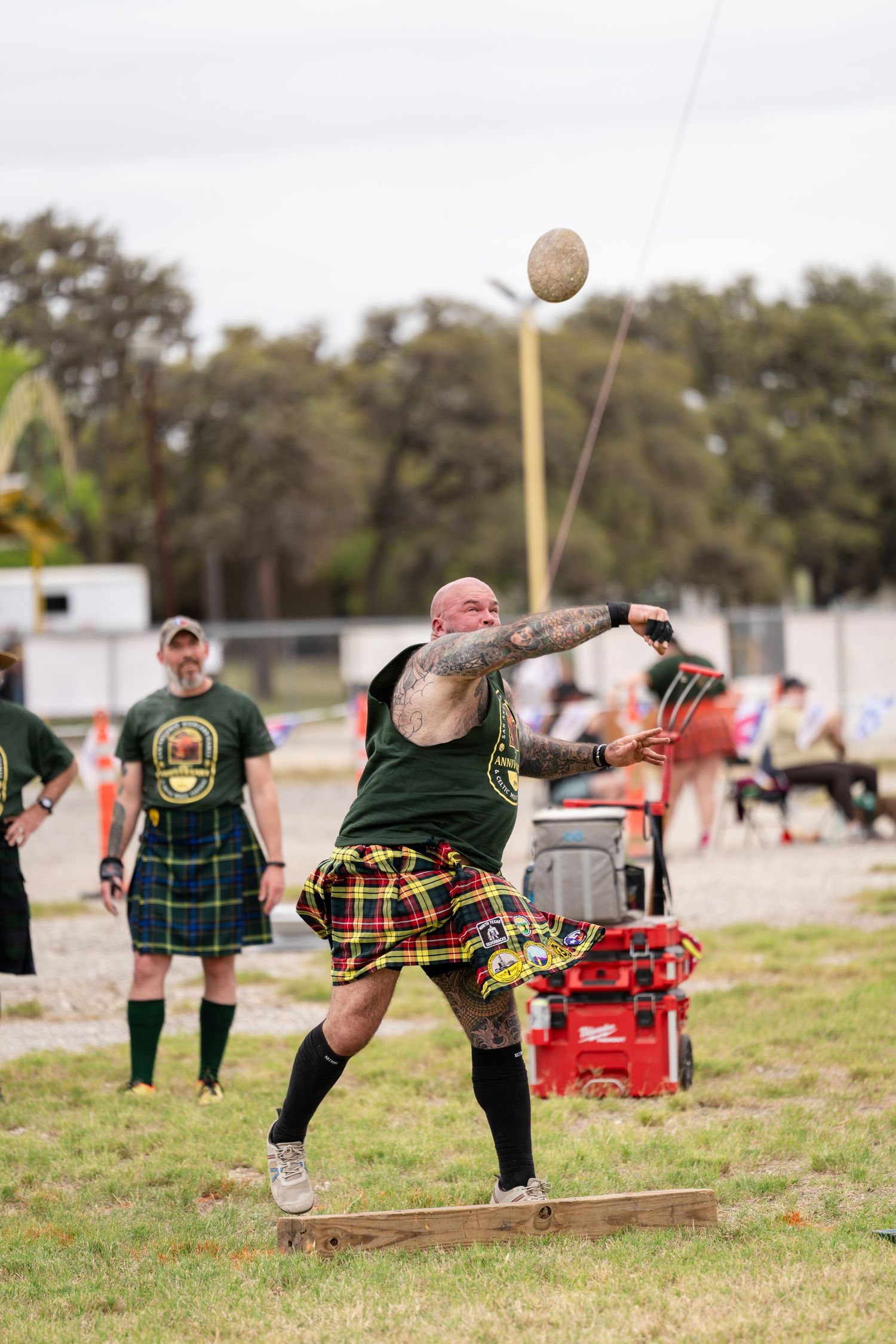 Tattooed athlete in tartan kilt throws heavy stone at Scottish Highland Games competition outdoors.