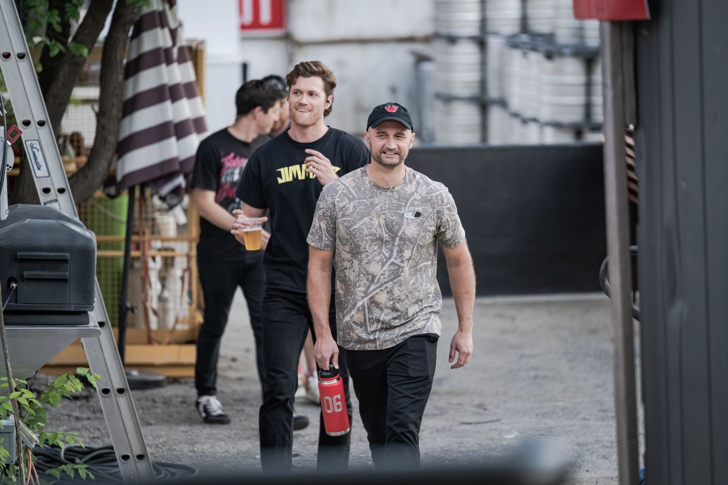 Three men walking outdoors near a building, one holding a red can and another carrying a beer glass.