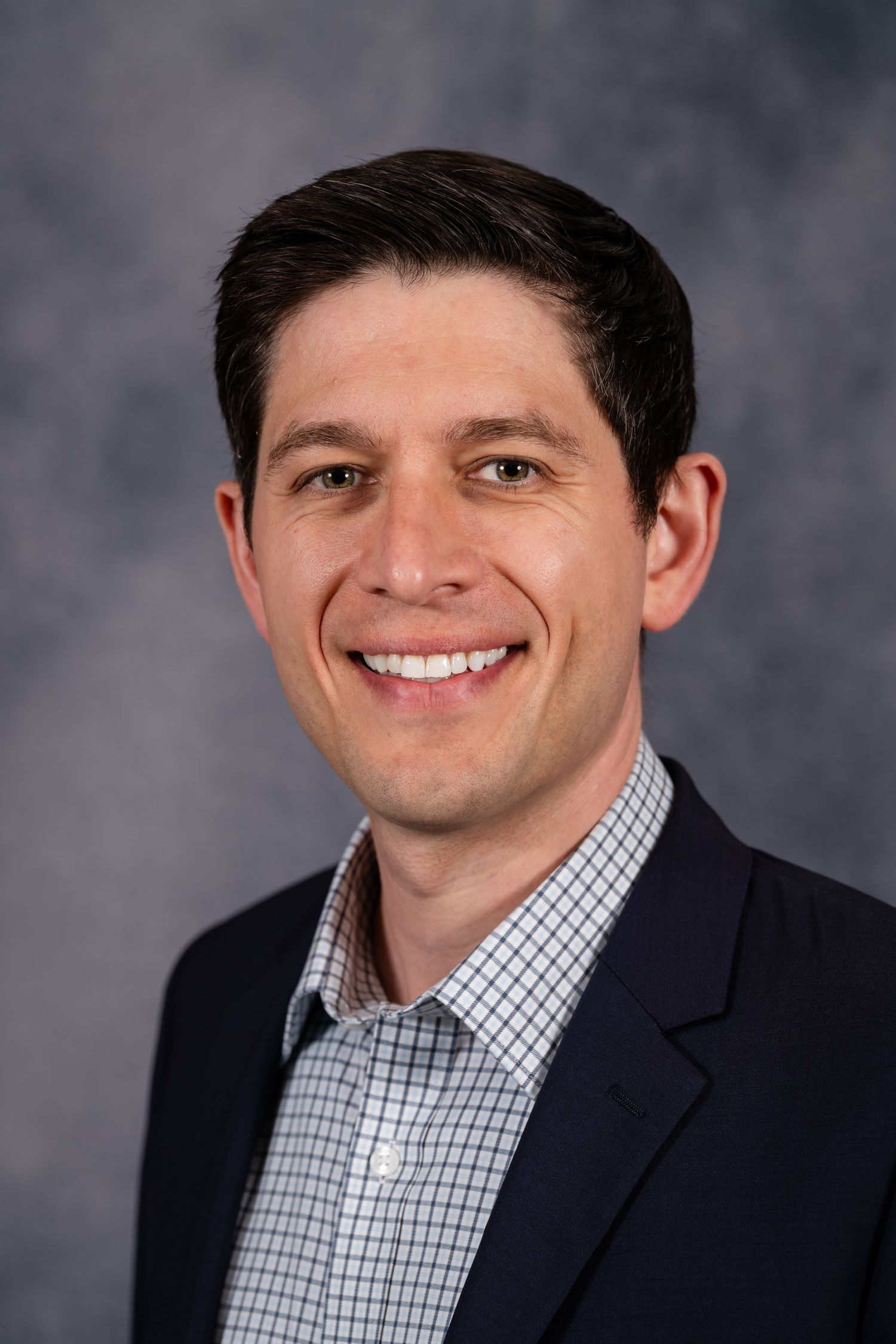 Professional headshot of a smiling young man in a navy blazer and checkered shirt against a gray background.