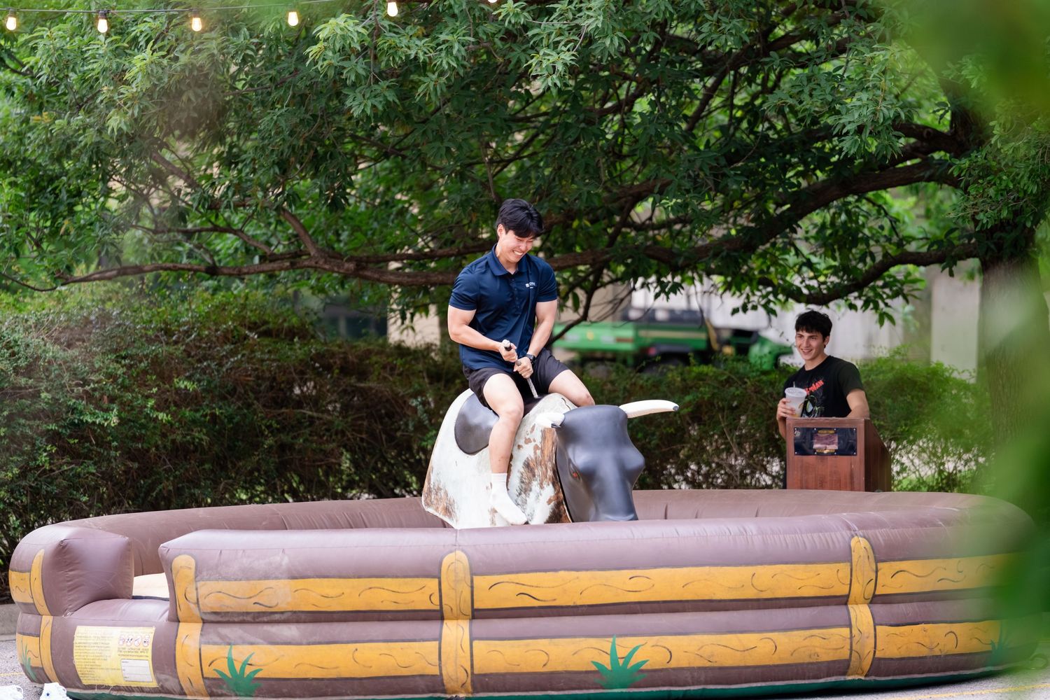 Man riding mechanical bull in inflatable arena outdoors, surrounded by lush green trees at a fun outdoor event.