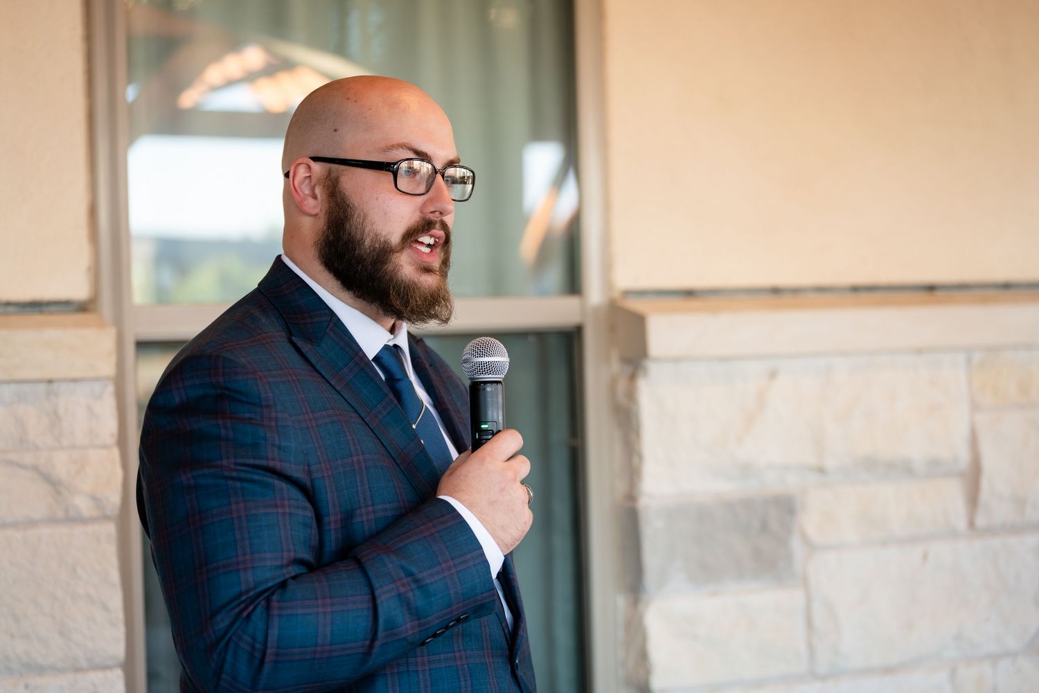 A person in a navy checkered suit and glasses speaks into a microphone against a beige stone wall background.