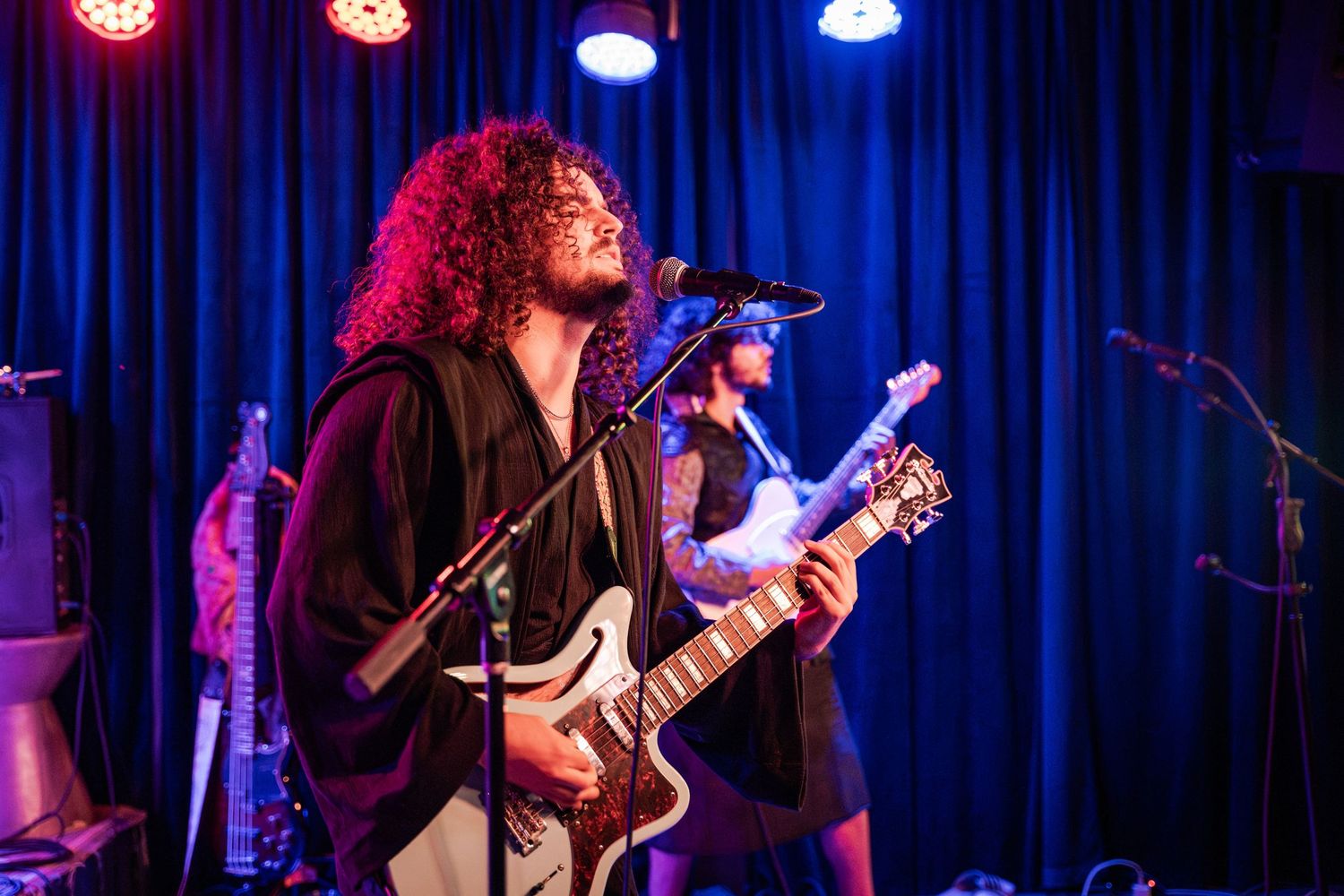 A musician performs on stage with a guitar under dramatic blue and red stage lighting during a live concert performance.