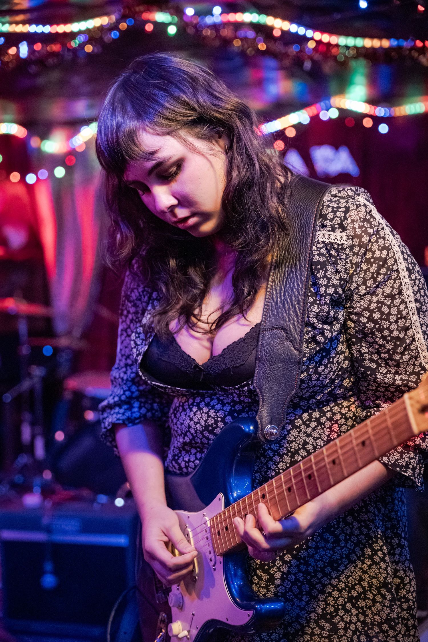 Musician in a patterned dress plays electric guitar on dimly lit stage with colorful bokeh lights in background.
