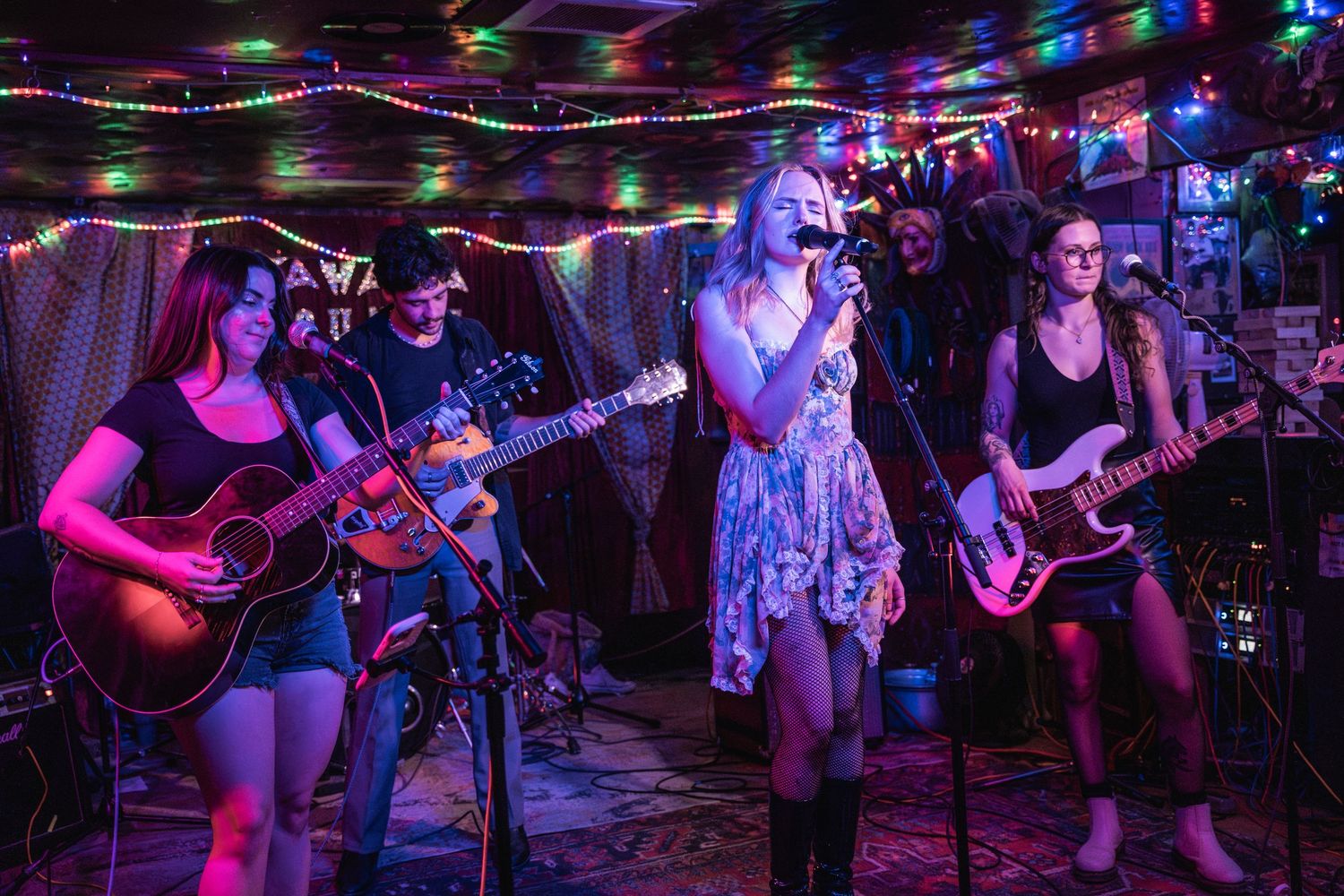 A live band performs on stage with colorful lighting, featuring musicians playing guitar and singing at a small venue.