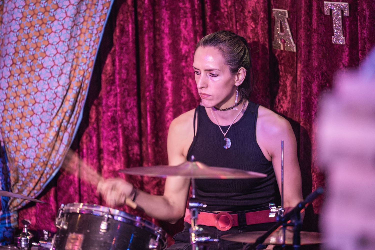 A drummer performs on stage with red velvet curtains in the background during a live music performance.