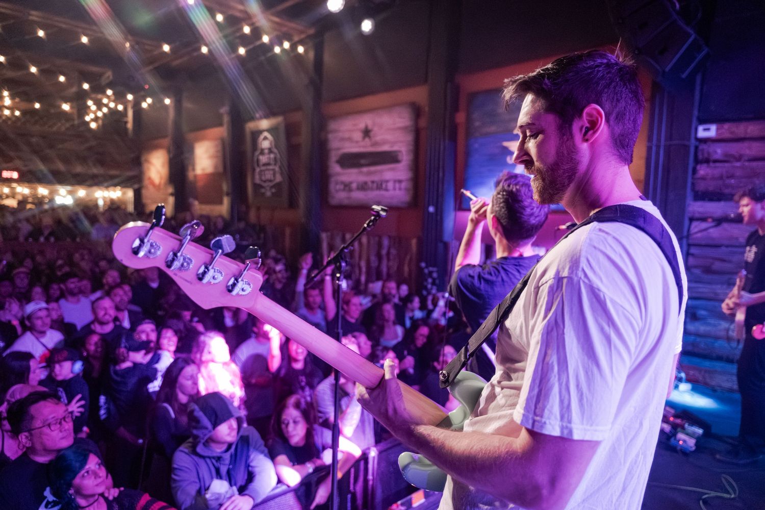 A band performs on stage in a dimly lit venue with colorful purple lighting and an energetic crowd.