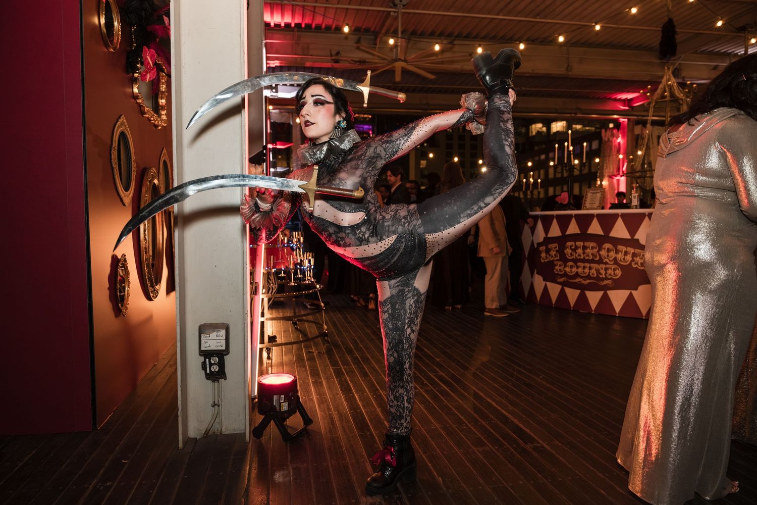 Dancer performs dramatic high kick pose in dark atmospheric venue with red lighting and wooden floors.
