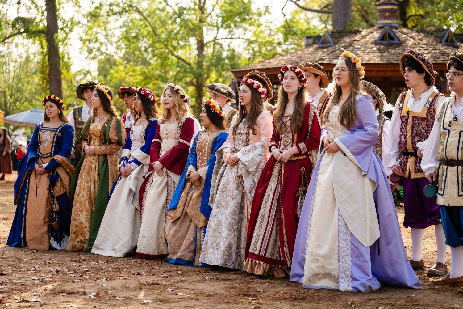 A group in Renaissance period costumes poses together at an outdoor festival wearing elaborate gowns and historical attire.