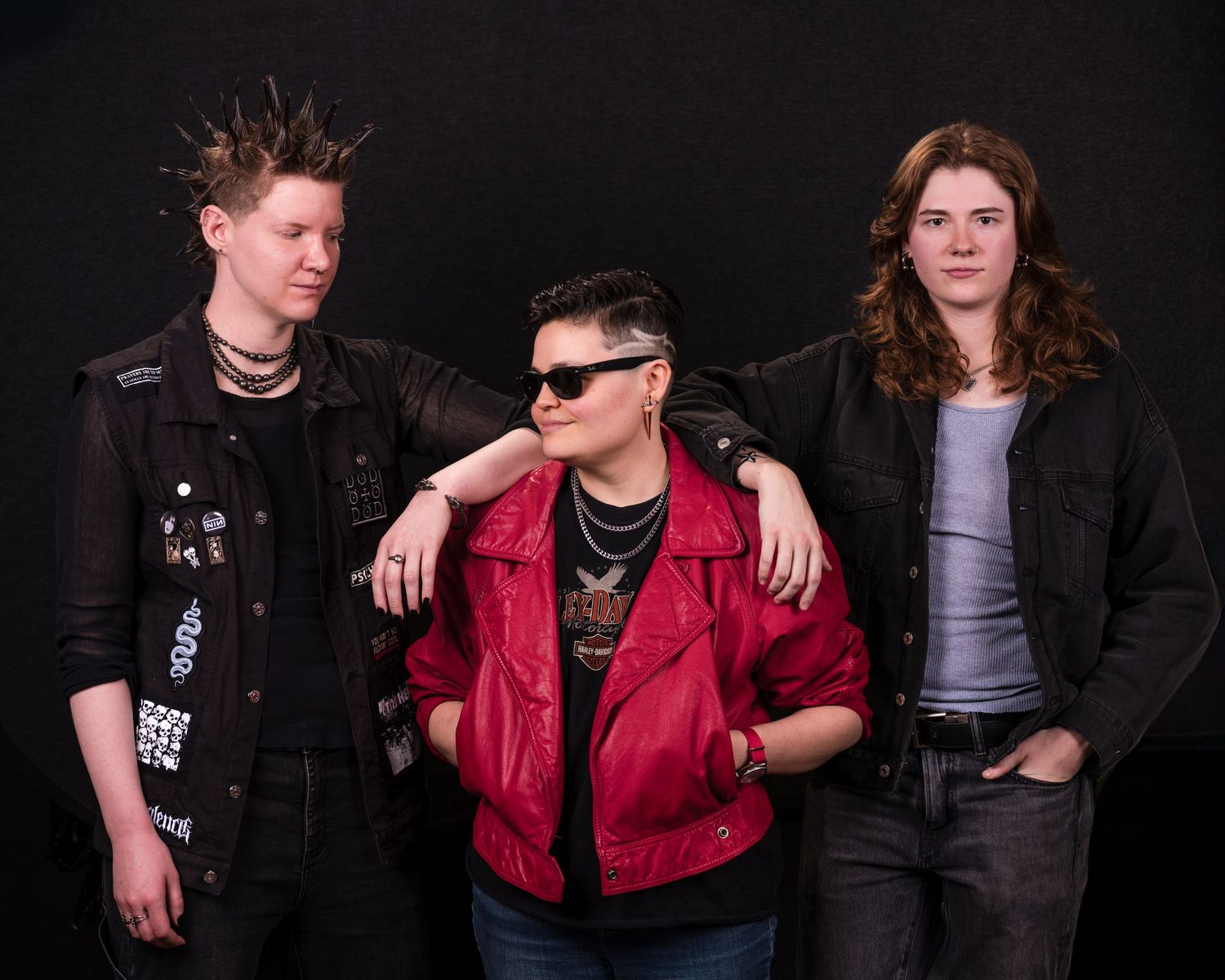 Three musicians pose together in a studio portrait with one wearing a bright red jacket and sunglasses.