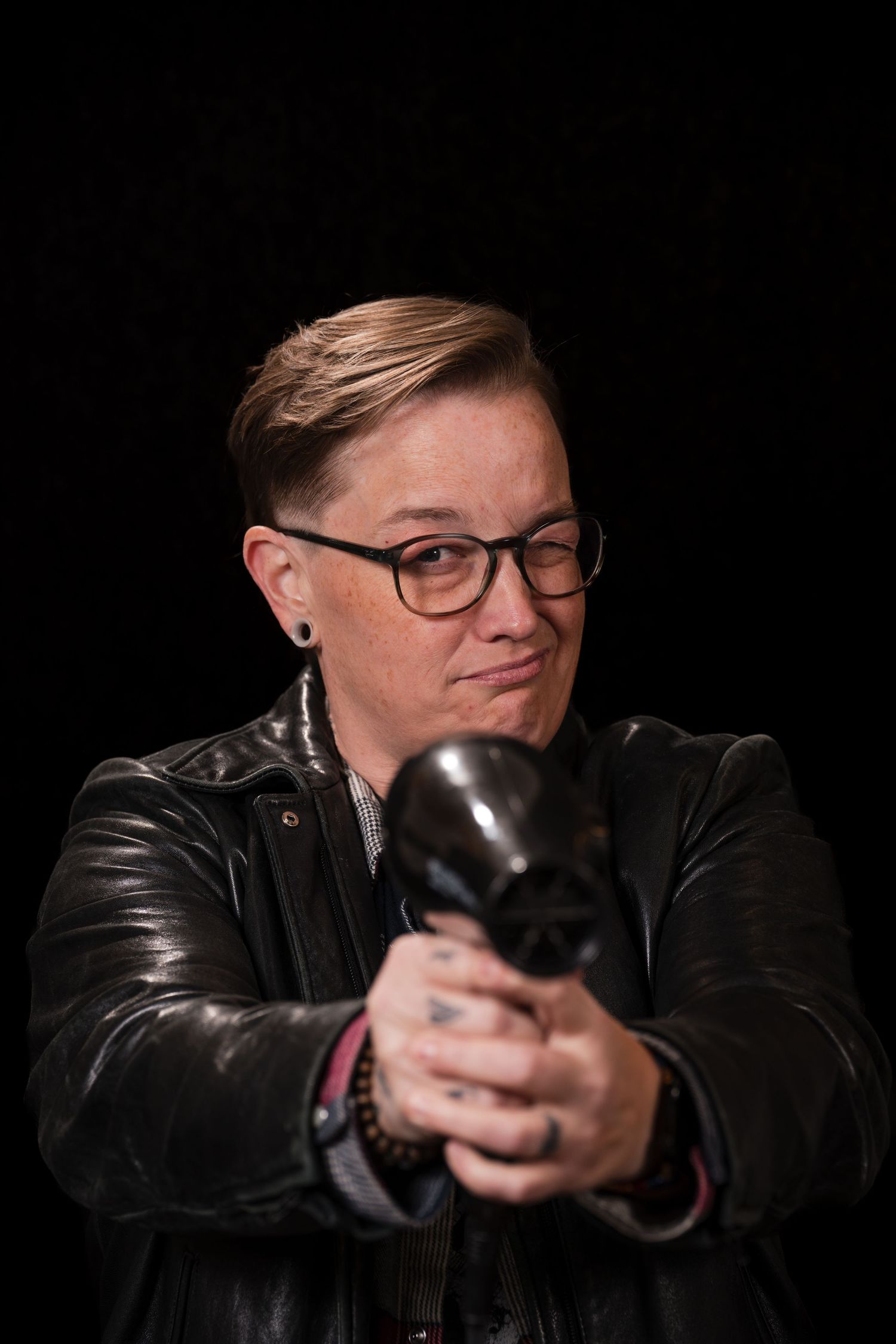 Person in leather jacket and glasses holds a dark object against black background in dramatic lighting.