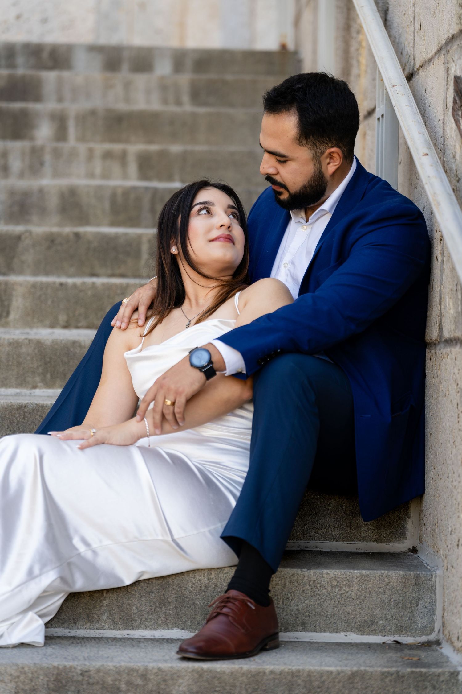 A couple in formal attire sits intimately on concrete stairs, with her in white and him in a navy blue suit.