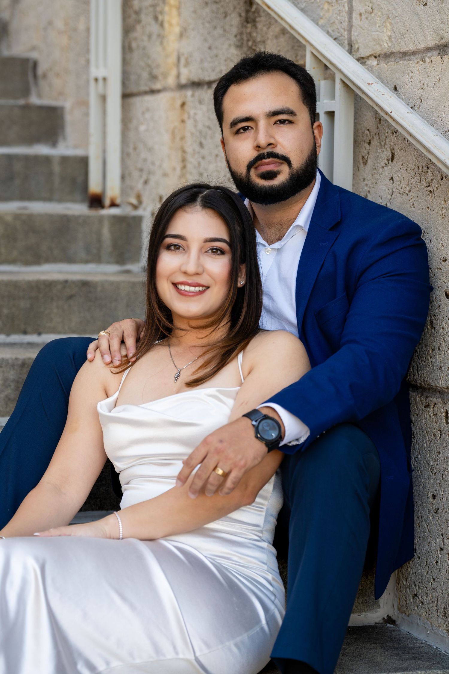 Couple in formal attire sitting on concrete stairs, with woman in white dress and companion in navy blue suit.