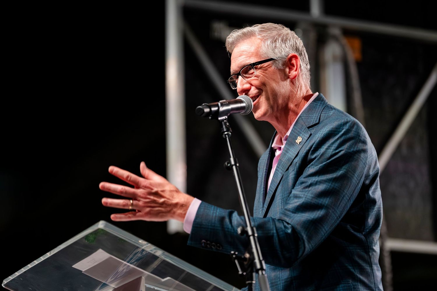 A person in a dark suit gestures expressively while speaking at a podium during a presentation.