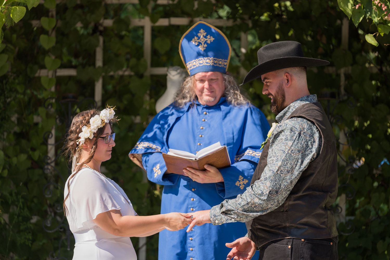 A wedding ceremony takes place outdoors with people gathered in formal attire under green foliage.