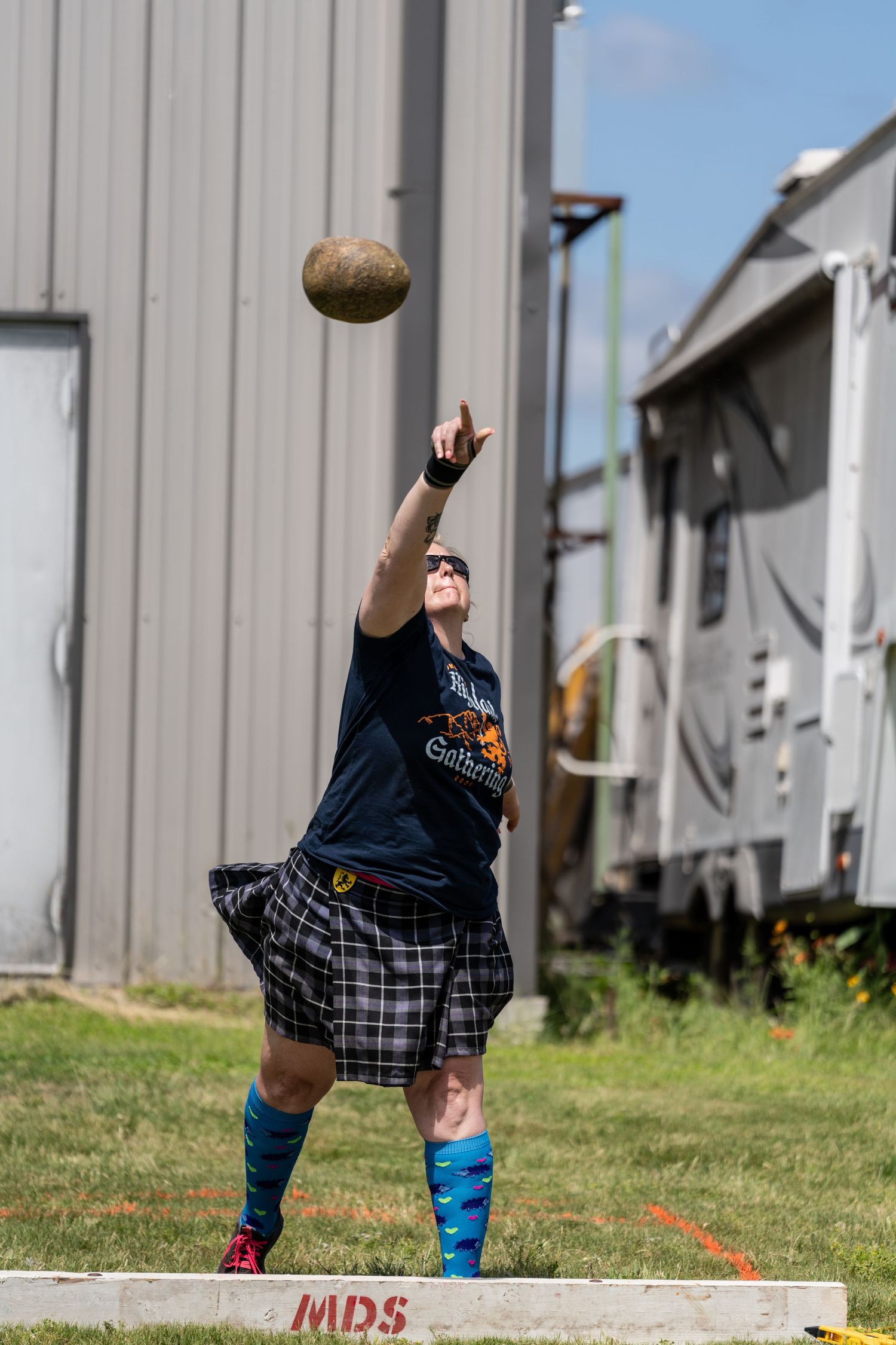 Man in kilt throwing stone ball at Highland Games competition outdoors near metal buildings.