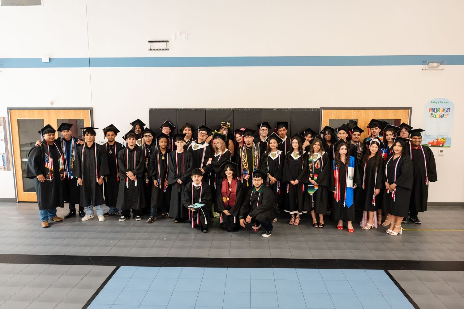 Large group of graduates in caps and gowns posing together in a gymnasium for a graduation photo.