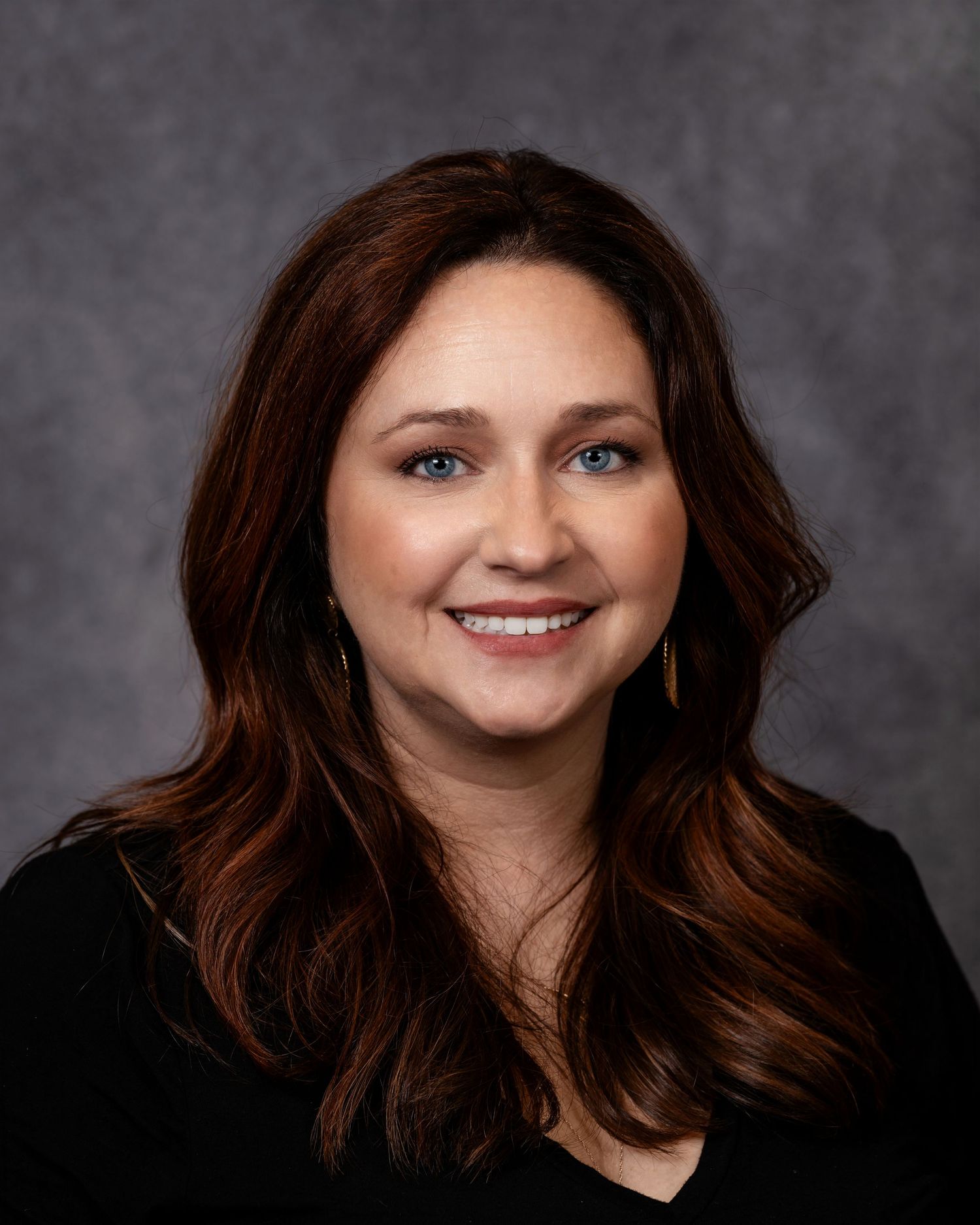 Professional headshot of a smiling woman with long auburn hair wearing a black top and gold earrings.