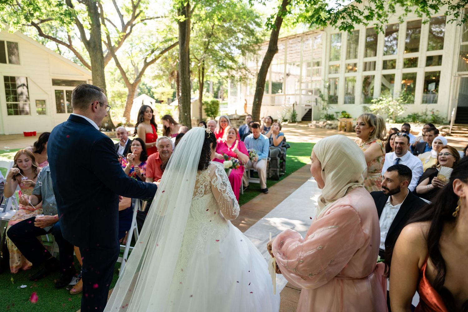 Outdoor wedding ceremony with bride in white gown and groom at altar, surrounded by seated guests in a sunlit garden.