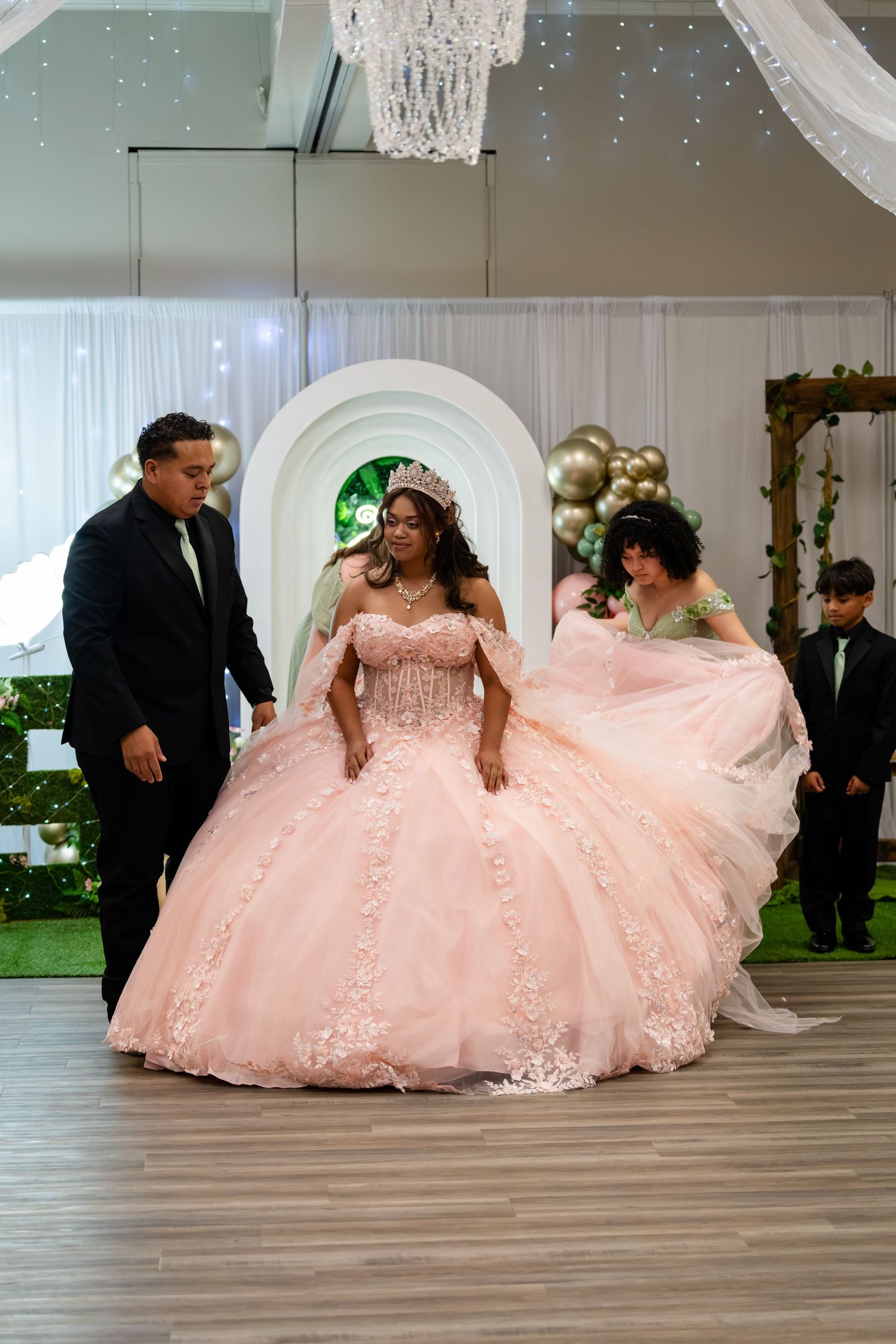 Young woman in pink quinceañera ballgown with tiara celebrates her quinceañera surrounded by family in decorated venue.