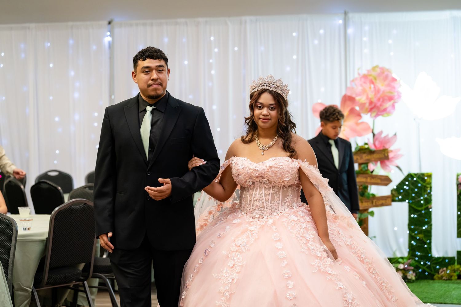 Young couple at a quinceañera, girl in pink ball gown with tiara, escort in black suit, elegant venue.