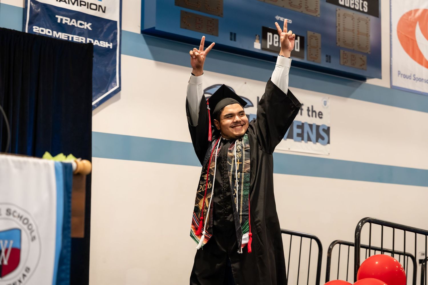 Graduate in cap and gown celebrates at graduation ceremony, raising hands in victory pose inside school gymnasium.