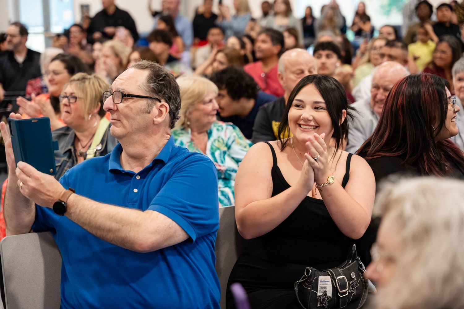 Audience members clapping and smiling at an indoor event, with a crowd of diverse attendees visible in the background.