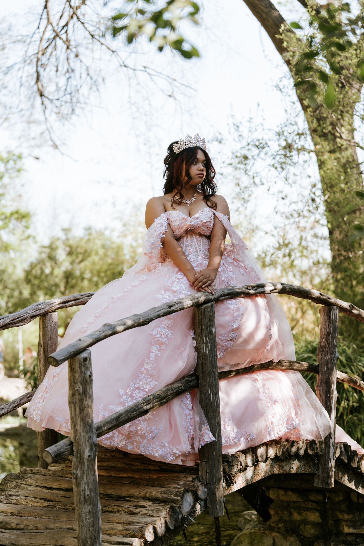 Young woman in pink quinceañera ball gown with lace details and tiara poses on rustic wooden bridge outdoors.
