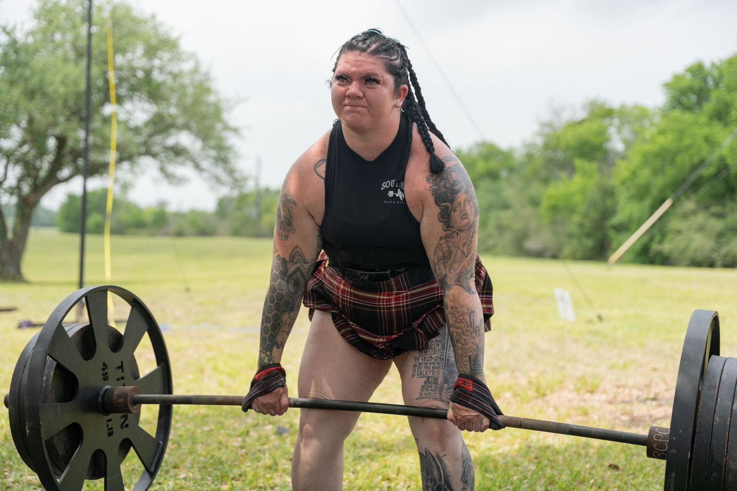 Tattooed strongman competitor deadlifting heavy barbell outdoors at Highland Games competition wearing kilt.