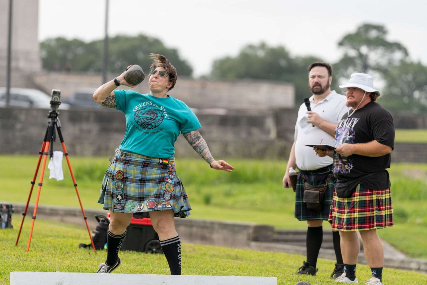 Athletes in kilts compete in Highland Games shot put event outdoors, with judges observing nearby.