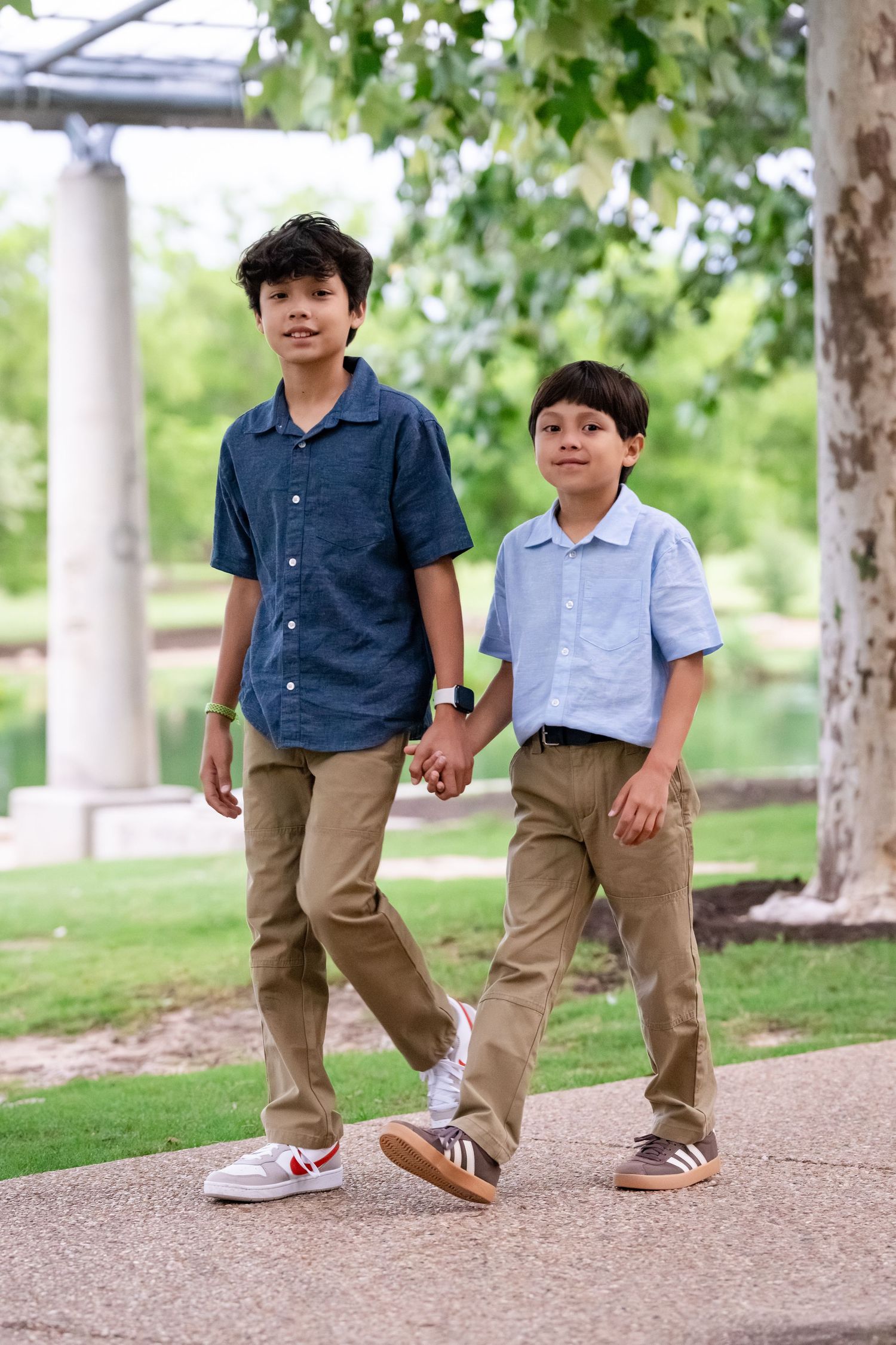Two boys holding hands walk along a park path, wearing button-up shirts and khaki pants on a sunny day.