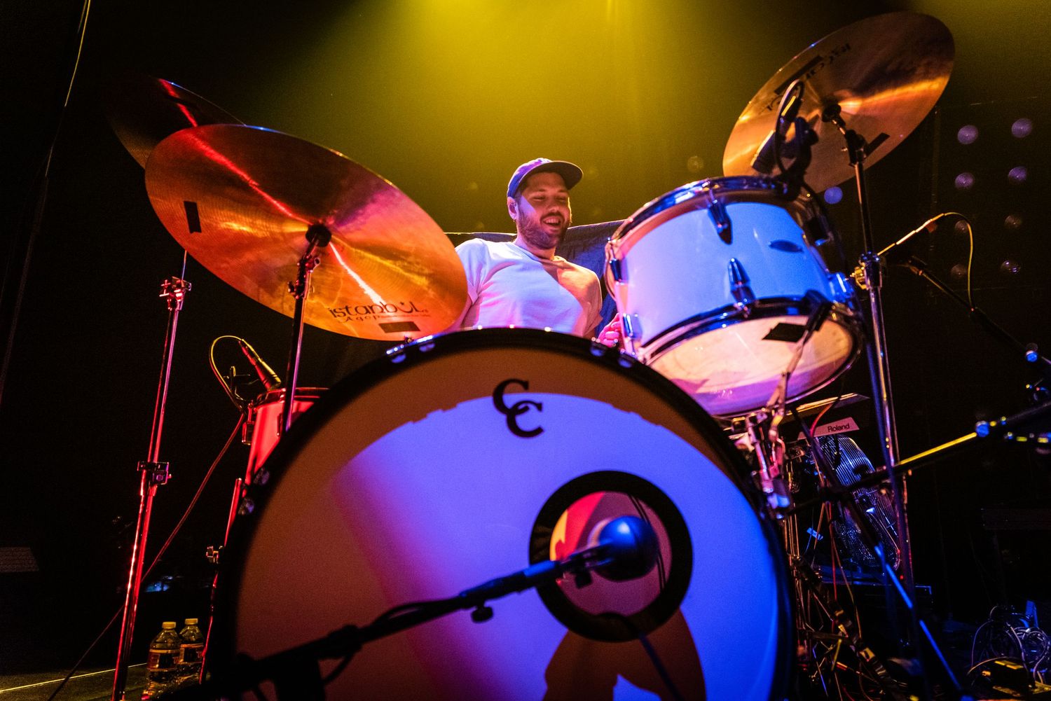 Drummer performs behind illuminated drum kit with bright yellow stage lighting.