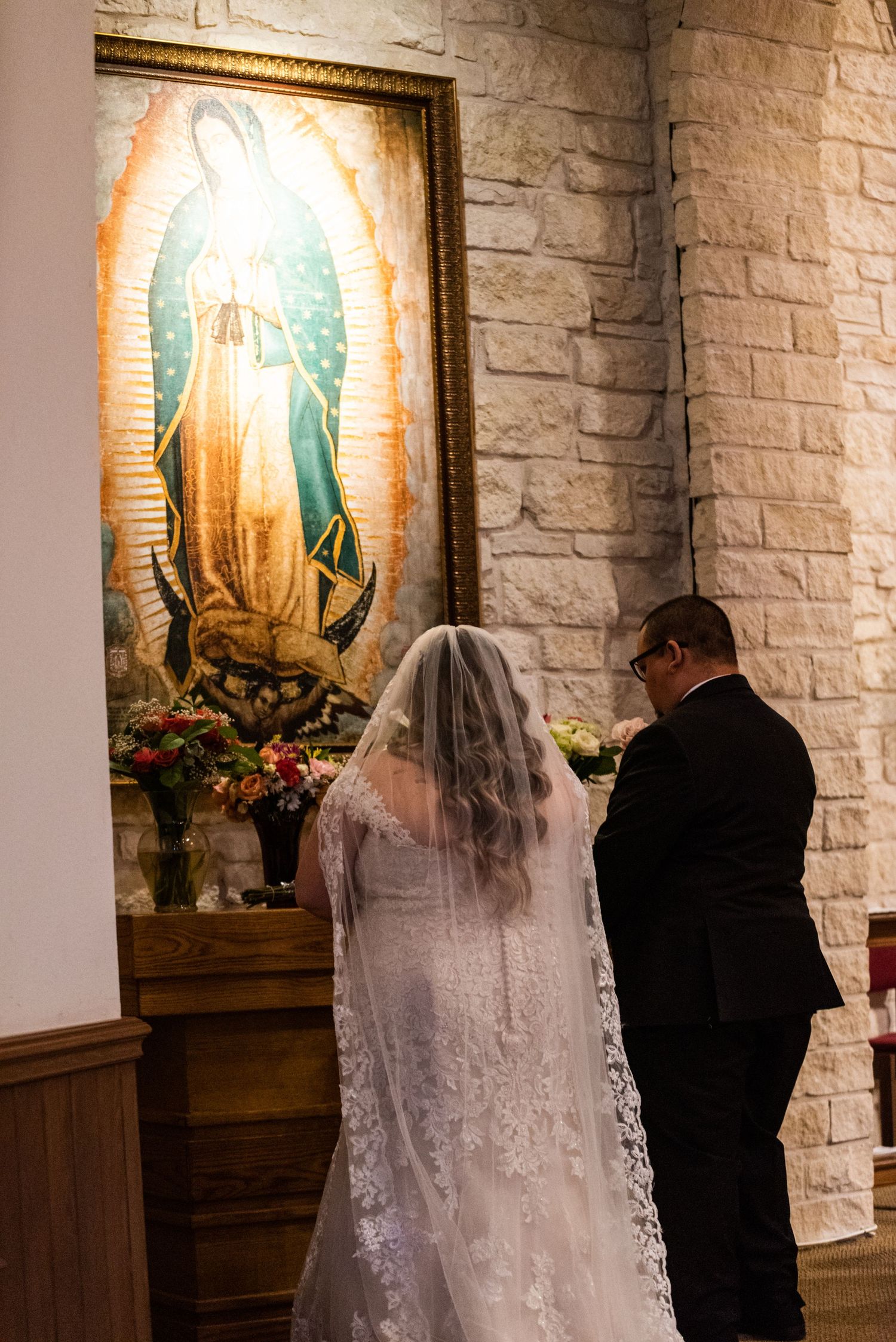 A couple prays before a painting of Our Lady of Guadalupe in a stone church.
