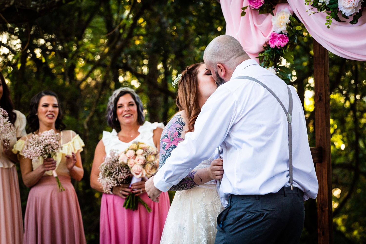 Outdoor wedding ceremony with pink drapery and floral arrangements under tree canopy.