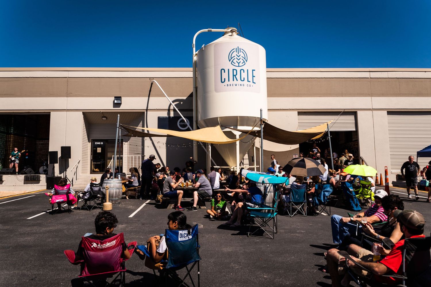 Crowd gathered under shade structures near large brewing tank at outdoor event.
