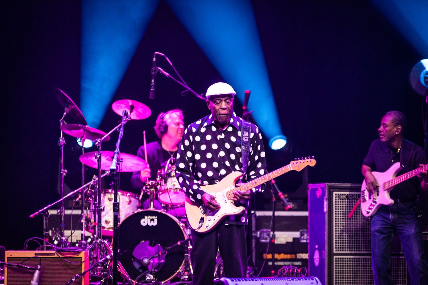 Guitarist in black and white polka dot shirt plays with full band on illuminated stage.