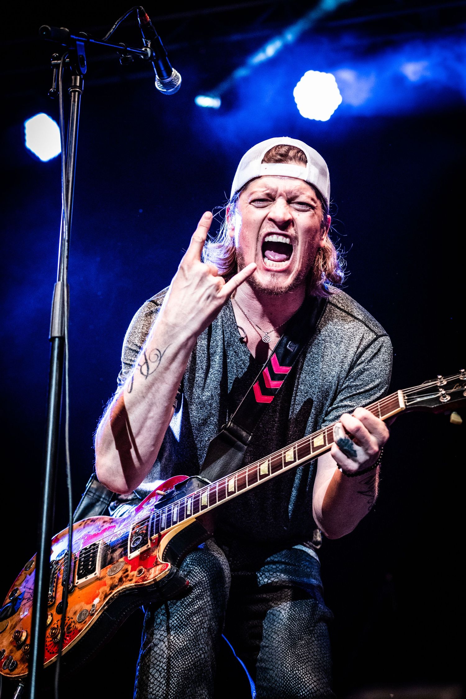 Energetic rock guitarist in white cap and gray jacket performs passionately on stage with blue lighting.